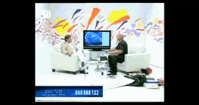 a man sits in a chair talking to another man in front of a tv screen