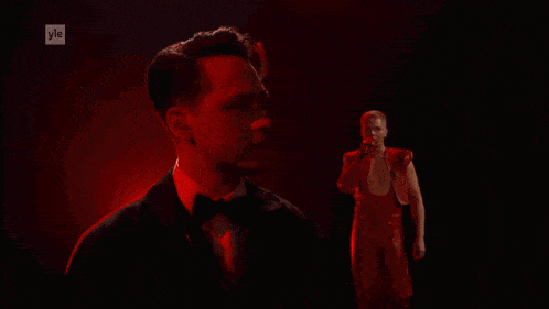 a man in a tuxedo holds a microphone while another man in a red shirt sings into it
