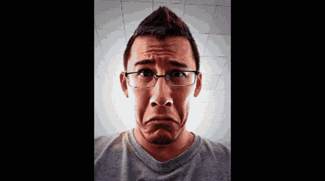 a man wearing glasses and a grey shirt is making a sad face