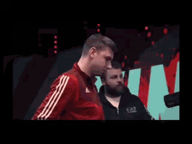 a man in a red adidas jacket is standing next to another man in a black ea7 jacket .