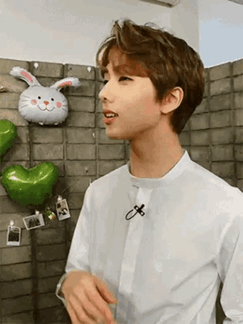a young man in a white shirt is standing in front of a brick wall with balloons and a stuffed rabbit .