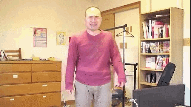a bald man in a pink shirt is standing in a room with a dresser and bookshelf .