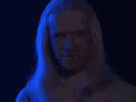 a man with long blonde hair is standing in a dark room with blue lights behind him .
