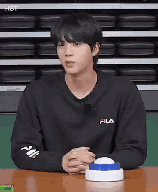 a young man in a black sweatshirt is sitting at a table with his hands folded .