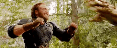 a man with a beard is standing in the woods holding a sword in his hand .