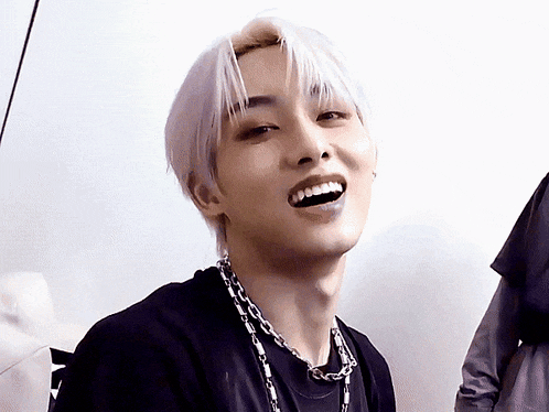 a young man with white hair is smiling and wearing a black shirt and a chain necklace .