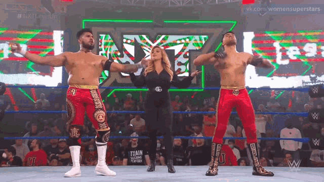 a woman stands between two men in a wrestling ring with a sign that says nxt on it