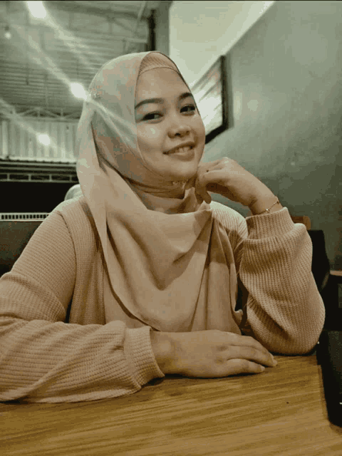 a woman wearing a hijab sits at a table with her hands on her chin