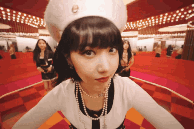 a woman wearing a pearl necklace and a white hat looks at the camera