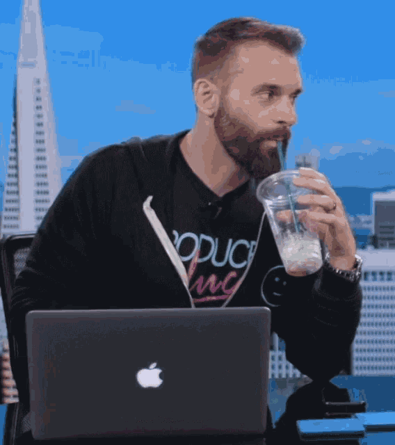 a man with a beard is drinking from a plastic cup with a straw in front of an apple laptop