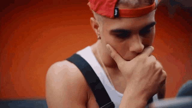 a young man wearing a red hat and a white tank top is holding his hand to his chin .