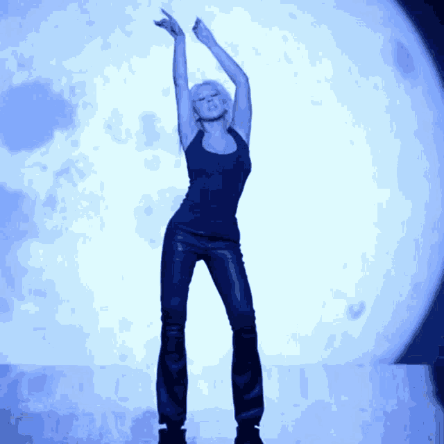 a woman stands in front of a blue moon with her arms in the air
