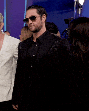a man wearing sunglasses and a black suit stands in a crowd of people