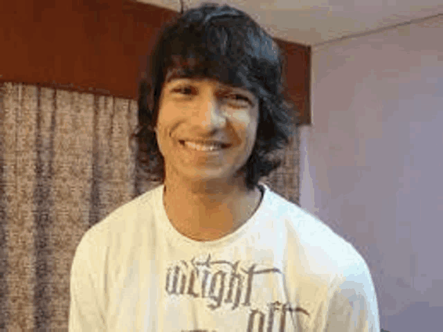 a young man with long hair is wearing a white t-shirt and smiling for the camera .
