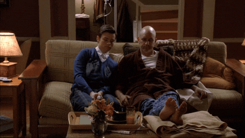 two men laying on a couch with a tray of food on the coffee table
