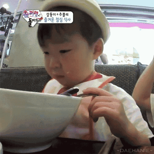 a little boy is eating from a bowl with a spoon while wearing a hat and bib ..