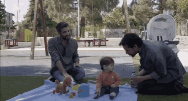 two men are playing with a baby on a blanket in a park and the baby is wearing an orange shirt