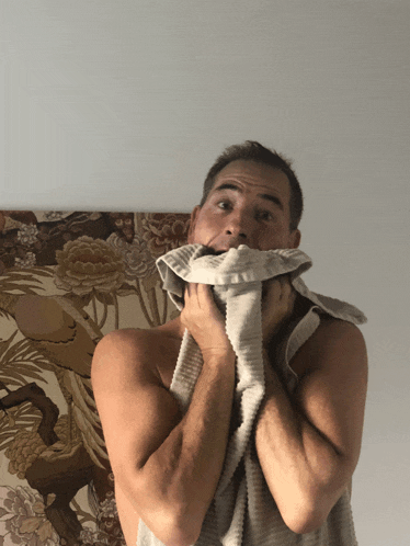 a shirtless man covering his face with a towel in front of a floral wallpaper