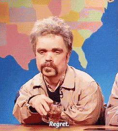 a man with a beard is sitting at a table holding a glass of whiskey and saying " regret "