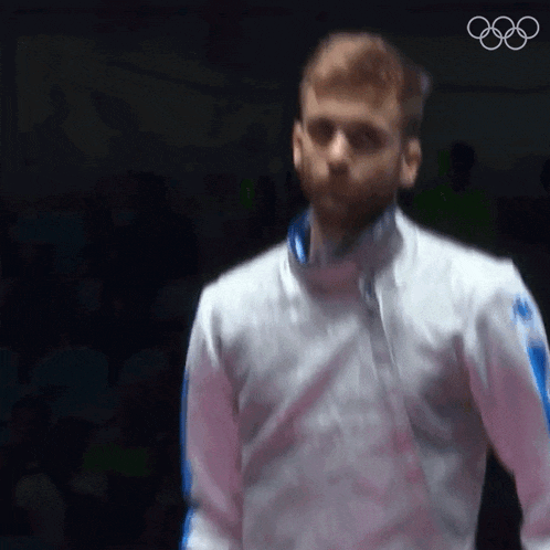 a man in a white jacket is standing in front of a black background with the olympic rings in the background .