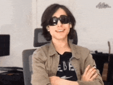 a young man wearing sunglasses is sitting in a chair with his arms crossed and smiling .