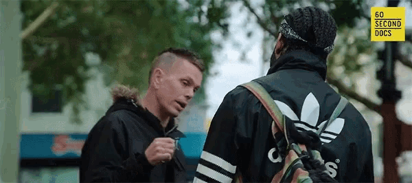 a man in an adidas jacket is talking to another man in a black jacket .