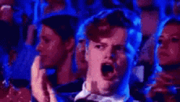 a man is screaming in a crowd of people in a dark room with blue lights behind him .