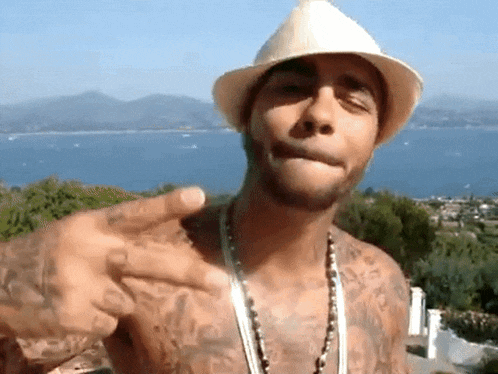 a shirtless man wearing a hat and a necklace is pointing at something
