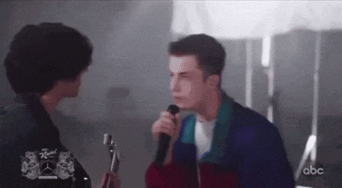 a man is singing into a microphone while another man plays a guitar in the background .