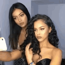 two women are taking a selfie with a cell phone . one of the women is wearing a black dress .
