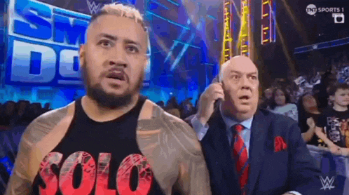 two men are standing next to each other in a wrestling ring . one of the men is talking on a cell phone .