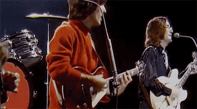 a man in a red sweater is playing a guitar in front of a drum set .