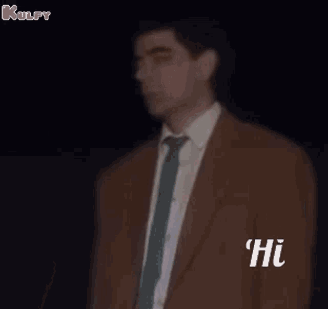 a man in a suit and tie is standing in the dark and saying hi .