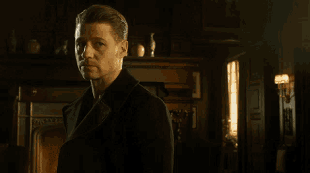 a man in a black coat stands in front of a fireplace in a dark room