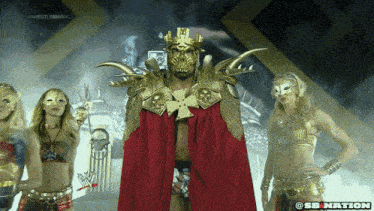 a man in a gold and red costume is surrounded by women in masks and the words wrestlemania are on the screen behind him