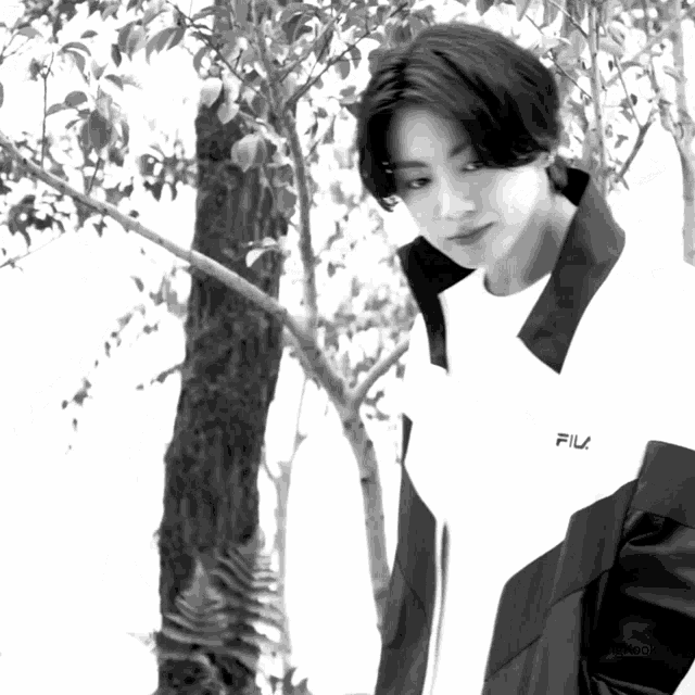 a black and white photo of a man wearing a fila jacket standing next to a tree