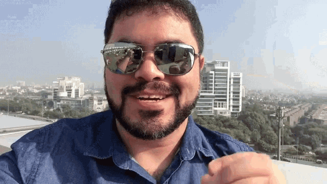 a man with a beard wearing sunglasses and a blue shirt with a city in the background