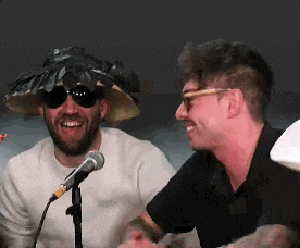 a man wearing a hat and sunglasses is talking into a microphone while another man smiles .
