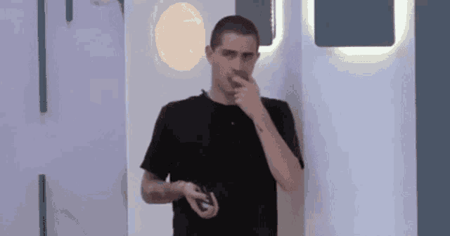 a man in a black shirt is standing in front of a wall and covering his nose with his hand .
