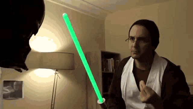 a man is holding a green light saber in front of another man in a dark helmet