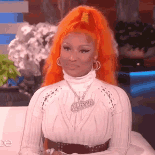 a woman with bright orange hair is wearing a white sweater and a necklace with the word queen on it