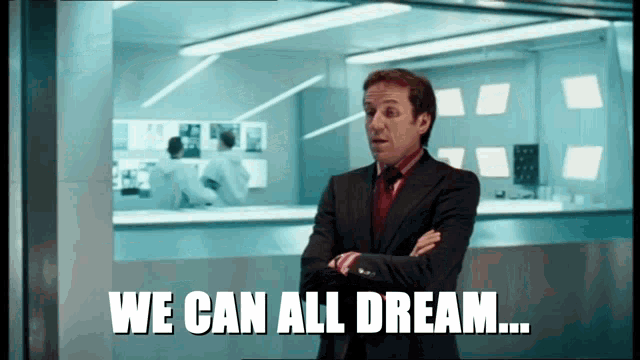 a man in a suit and red tie is standing in front of a window and says we can all dream