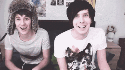 two young men are sitting next to each other on a bed and smiling . one of the men is wearing a fur hat .