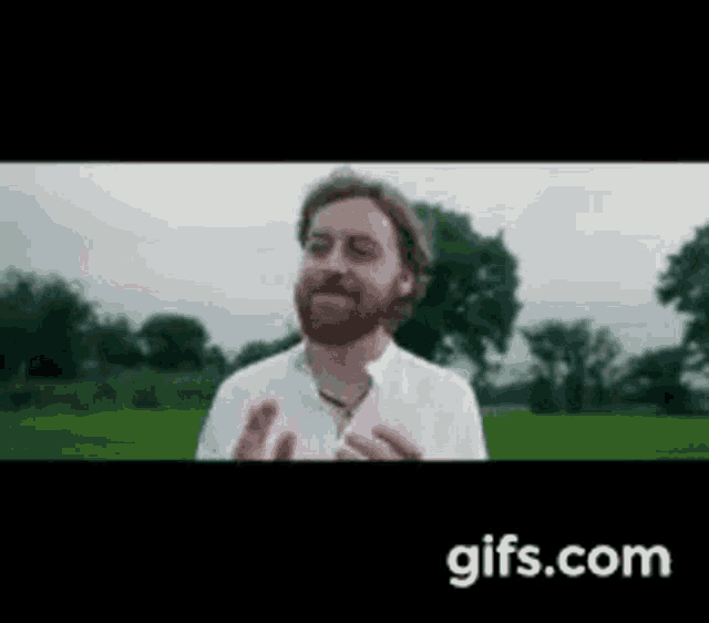 a man with a beard is standing in a field with trees in the background and talking to someone .
