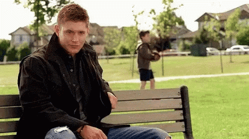 a man is sitting on a bench in a park with a boy standing behind him .