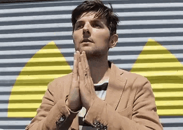 a man in a tan jacket is praying with his hands folded in front of a yellow and gray wall .