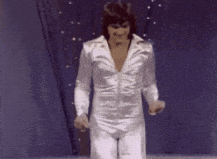 a man in a white jumpsuit is dancing on a stage in front of a blue curtain .