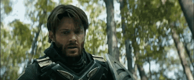 a man with a beard is standing in the woods with a sword .