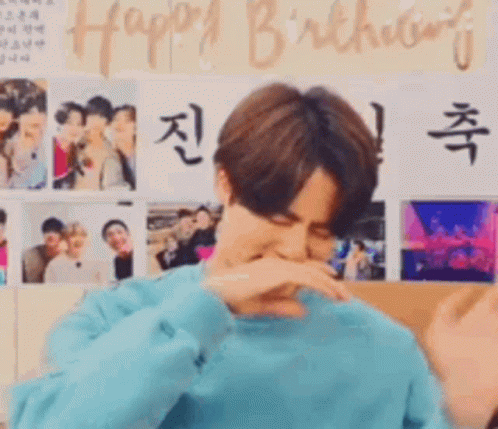 a man in a blue sweater is covering his mouth with his hand in front of a wall with pictures and the words happy birthday .