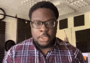 a man wearing glasses and a plaid shirt stands in front of a wall with a clock on it